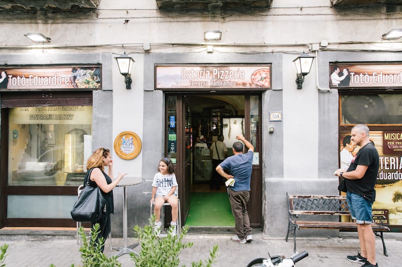 NAPLES, ITALY - 13 JULY 2019: Customers are seen here at the entrance of the Osteria Totò Eduardo e Pasta e Fagioli, a restaurant in Naples, Italy, on July 13th 2019.<br /> The name of the Osteria is dedicated to the two masters of Neapolitan theatre and cinema: Totò (Antonio de Curtis) and Eduardo de Filippo. The idea came from Mario Bianchini, an aficionado of Neapolitan culture who wanted to pay tribute to his wife Rosaria de Curtis, a distant relative of Totò, the most famous actor in the history of Naples.<br /> The osteria was founded in the 1970s by Mario Bianchini who wanted to base its menu on traditional Neapolitan dishes. Mr Bianchini learned the art of cooking from his mother Anna.