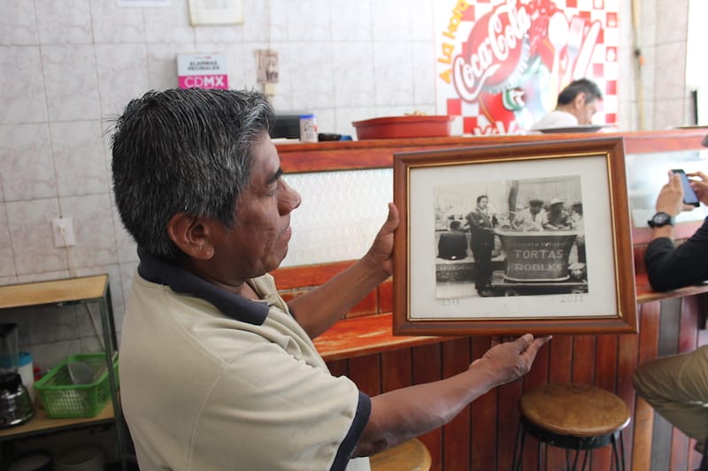 A server holds up a photo of Alejandro Robles, with his torta cart in 1947, before he rented a storefront, photo by Martha Pskowski