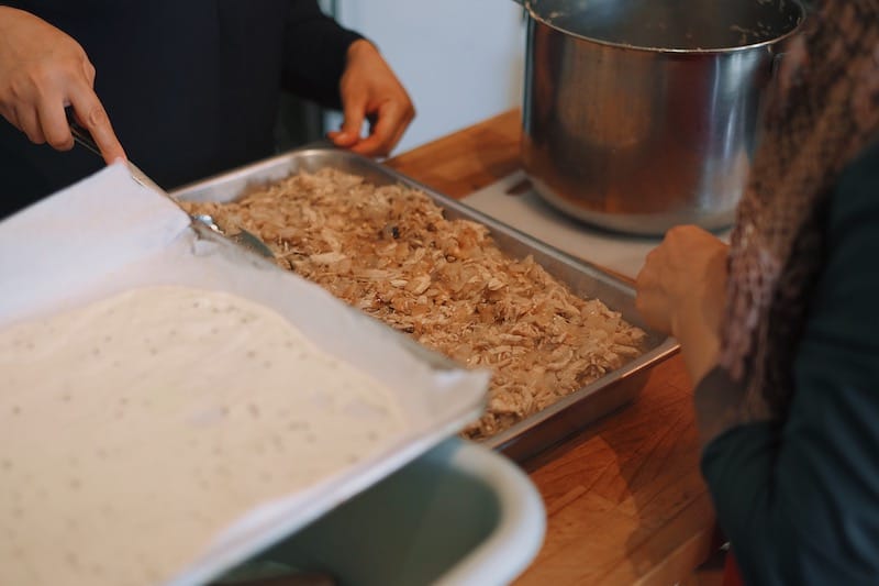 Two Syrian women prepare el r’gaga, poached chicken and caramelized onions baked in layers of rolled dough, at Newcomer Kitchen, photo by Mandy Sham newcomer kitchen toronto
